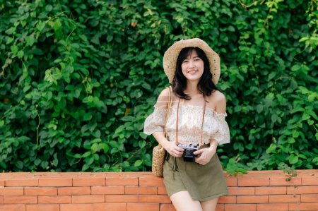 Photo for Portrait of young asian woman traveler with weaving hat and basket and a camera on green public park nature background. Journey trip lifestyle, world travel explorer or Asia summer tourism concept. - Royalty Free Image