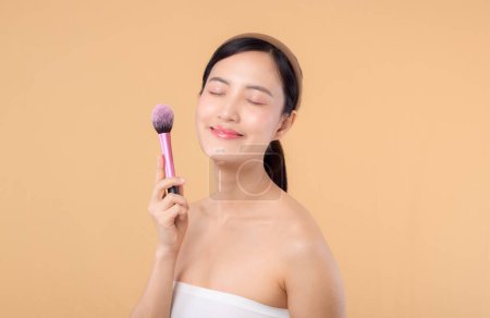 Téléchargez les photos : Portrait jeune femme asiatique heureuse avec naturel maquillage visage tenant peau cosmétique poudre rougisseur isolé sur fond beige. Femme appliquer un traitement de brosse de soins de la peau. produit de beauté, concept de cosmétologie - en image libre de droit