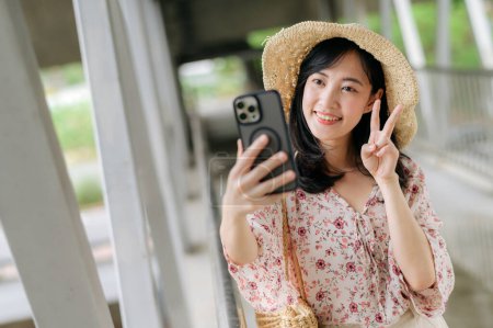 Photo for Asian young woman traveler with weaving basket selfie with mobile phone and standing on overpass. Journey trip lifestyle, world travel explorer or Asia summer tourism concept. - Royalty Free Image