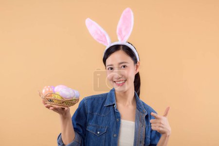 Photo for Portrait asian young woman wearing denim clothes and bunny rabbit ears and holding colorful eggs isolated on beige background. Lifestyle Happy Easter concept. - Royalty Free Image