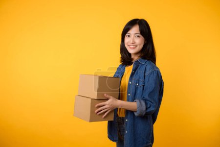 Foto de Retrato de mujer asiática joven con camiseta amarilla y camisa de mezclilla que sostiene la caja de paquetes aislada en el fondo del estudio amarillo, mensajero de entrega y concepto de servicio de envío. - Imagen libre de derechos