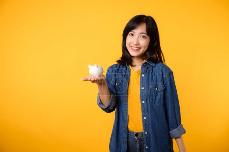 Téléchargez les photos : Portrait jeune femme asiatique gaie portant t-shirt jaune chemise en denim avec sourire heureux tenant tirelire économiser de l'argent d'investissement isolé sur fond jaune. économie finance entreprise richesse concept. - en image libre de droit
