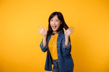 Photo for Asian young woman wearing yellow t-shirt denim shirt with open mouths raising hands screaming announcement isolated on yellow background. Lifestyle portrait concept. - Royalty Free Image
