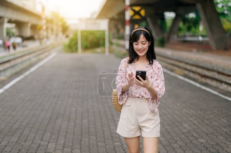 Foto de Joven mujer asiática viajero con cesta de tejido utilizando un teléfono móvil al lado de la estación de tren en Bangkok. Viaje viaje estilo de vida, viajero mundial o concepto de turismo de verano de Asia. - Imagen libre de derechos