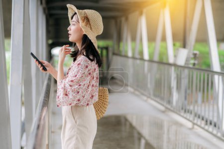 Foto de Asiático joven viajero con tejer cesta usando teléfono móvil y de pie en puente. Viaje viaje estilo de vida, viajero mundial o concepto de turismo de verano de Asia. - Imagen libre de derechos