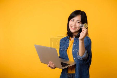 Photo for Happy asian young woman wearing yellow t-shirt denim shirt holding digital crypto currency coin and laptop isolated on yellow background. Digital currency financial concept. - Royalty Free Image