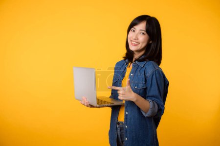 Photo for Portrait happy young woman wearing yellow t-shirt and denim shirt holding laptop and point finger to screen isolated on yellow studio background. business technology application communication concept. - Royalty Free Image