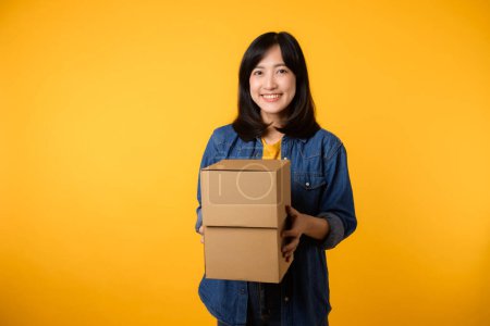 Téléchargez les photos : Portrait jeune femme asiatique portant t-shirt jaune et chemise en denim tenant boîte de colis isolé sur fond de studio jaune, livraison courrier et concept de service d'expédition. - en image libre de droit
