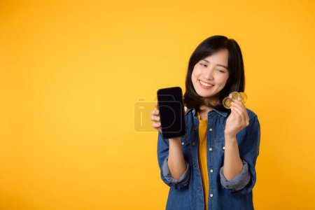 Foto de Feliz asiática joven mujer con camiseta amarilla camisa de mezclilla que sostiene moneda cripto digital moneda y teléfono inteligente aislado en el fondo amarillo. Concepto financiero de moneda digital. - Imagen libre de derechos
