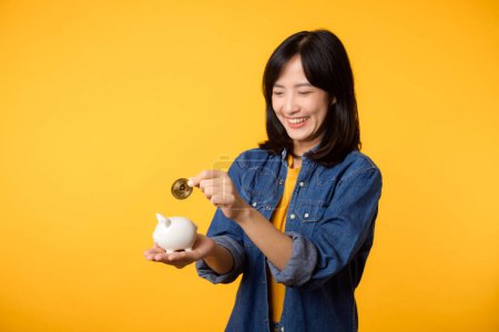 Foto de Mujer joven asiática feliz vistiendo camiseta amarilla camisa vaquera ahorrando criptomoneda digital a alcancía aislada sobre fondo amarillo. Concepto de inversión de ahorro digital. - Imagen libre de derechos