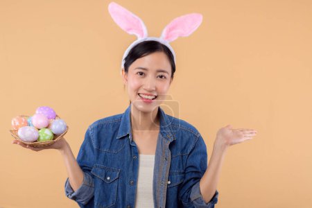 Téléchargez les photos : Portrait jeune femme asiatique portant des vêtements en denim et des oreilles de lapin et tenant des œufs colorés isolés sur fond beige. Lifestyle Joyeux concept de Pâques. - en image libre de droit