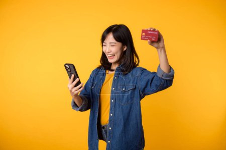 Foto de Retrato hermosa asiática joven sonrisa feliz vestido con chaqueta de mezclilla que muestra teléfono inteligente y tarjeta de crédito aislado en el fondo del estudio amarillo. Compras en línea smartphone concepto de aplicación - Imagen libre de derechos