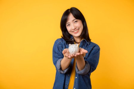 Téléchargez les photos : Portrait jeune femme asiatique gaie portant t-shirt jaune chemise en denim avec sourire heureux tenant tirelire économiser de l'argent d'investissement isolé sur fond jaune. économie finance entreprise richesse concept. - en image libre de droit