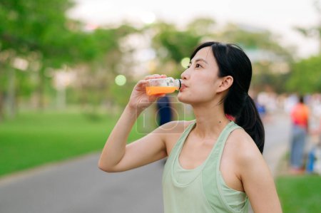 Foto de Una corredora. Ajuste mujer joven asiática con ropa deportiva verde beber jugo de naranja orgánico después de correr y disfrutar de un sano al aire libre. Chica corredor de fitness en el parque público. Bienestar siendo concepto - Imagen libre de derechos