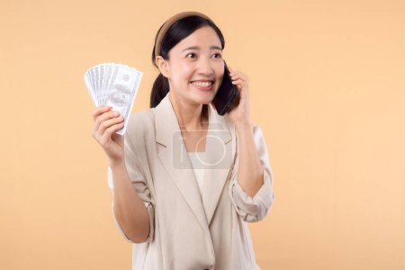 Téléchargez les photos : Portrait de heureux heureux confiant jeune femme d'affaires asiatique portant une veste blanche tenant smartphone et argent comptant dollar debout sur fond beige. millionnaire business, concept shopping. - en image libre de droit