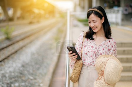 Foto de Joven viajera asiática con sombrero de tejer usando un teléfono móvil al lado de la estación de tren en Bangkok. Viaje viaje estilo de vida, viajero mundial o concepto de turismo de verano de Asia. - Imagen libre de derechos