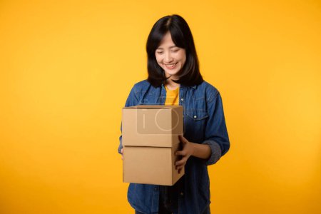 Téléchargez les photos : Portrait jeune femme asiatique portant t-shirt jaune et chemise en denim tenant boîte de colis isolé sur fond de studio jaune, livraison courrier et concept de service d'expédition. - en image libre de droit