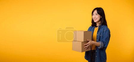 Photo for Portrait young asian woman wearing yellow t-shirt and denim shirt holding parcel box isolated on yellow studio background, Delivery courier and shipping service concept. - Royalty Free Image