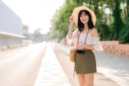 Photo for Portrait of young asian woman traveler with weaving hat and basket and a camera standing by the street. Journey trip lifestyle, world travel explorer or Asia summer tourism concept. - Royalty Free Image