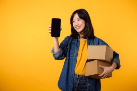 Photo for Portrait young asian woman wearing yellow t-shirt and denim shirt holding mobile phone and parcel box isolated on yellow studio background, Delivery courier and shipping service concept. - Royalty Free Image