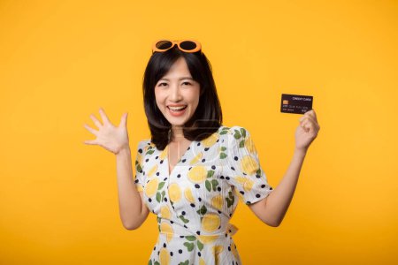 Foto de Retrato de mujer joven asiática feliz sonriendo en vestido de primavera mostrando tarjeta de crédito de plástico aislado sobre fondo amarillo. Pago, dinero y compra concepto de pago de compras. - Imagen libre de derechos
