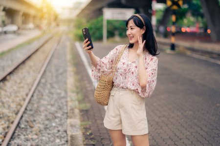 Photo for Young Asian woman traveler with weaving basket using a mobile phone beside railway train station in Bangkok. Journey trip lifestyle, world travel explorer or Asia summer tourism concept. - Royalty Free Image