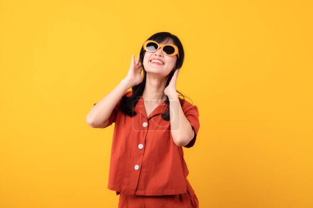 Foto de Retrato hermosa joven asiática mujer feliz sonrisa vestida con naranja ropa y gafas de sol mirando celebrar con la mano en su cara aislado en amarillo estudio fondo. - Imagen libre de derechos