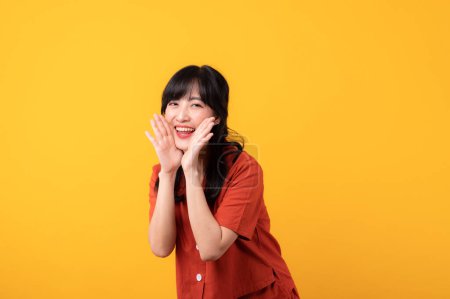Téléchargez les photos : Portrait belle jeune femme asiatique sourire heureux habillé de vêtements orange crier à l'espace libre isolé sur fond de studio jaune. Présentation du produit Belle fille agir comme dire au client - en image libre de droit