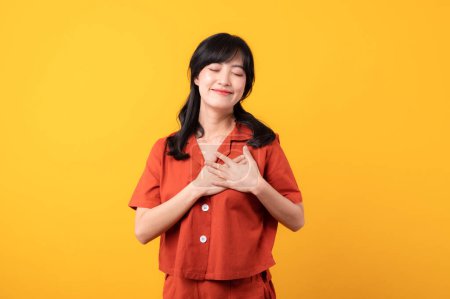 Téléchargez les photos : Portrait belle jeune femme asiatique sourire heureux habillé de vêtements orange montrant les mains touchant sa poitrine dans fier et reconnaissant sentiment émotionnel isolé sur fond jaune studio. - en image libre de droit