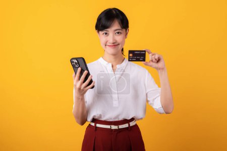 Foto de Retrato joven hermosa mujer asiática emprendedor sonrisa feliz con camisa blanca y plantas rojas que sostienen la tarjeta de crédito y el aislamiento de teléfonos inteligentes en el fondo amarillo. Concepto de compras online. - Imagen libre de derechos