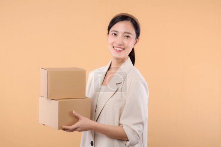 Photo for Portrait of successful happy confident young asian business woman wearing white jacket holding parcel box isolated on beige studio background, Delivery courier and shipping service concept. - Royalty Free Image