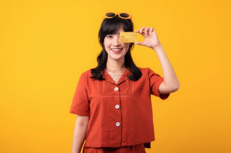Téléchargez les photos : Portrait belle jeune femme asiatique sourire heureux habillé de vêtements orange montrant carte de crédit isolé sur fond jaune. Paiement et achat shopping concept de paiement. - en image libre de droit