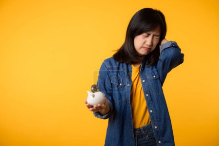 Photo for Unhappy young asian woman wearing yellow t-shirt denim shirt pulling digital coin crypto currency out of piggy bank isolated on yellow background. Payment digital money debt financial concept. - Royalty Free Image