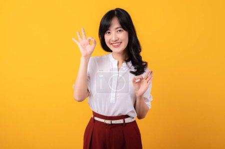 Foto de Retrato joven hermosa mujer asiática emprendedor feliz sonrisa usando camisa blanca y plantas rojas mostrando signo aceptable manos gesto aislado sobre fondo amarillo. - Imagen libre de derechos