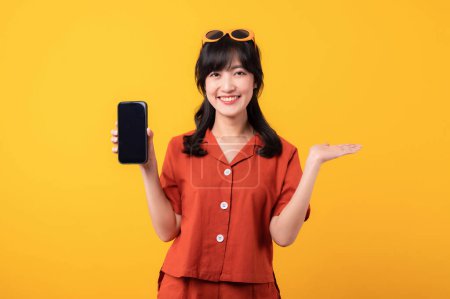 Téléchargez les photos : Portrait jeune belle femme asiatique sourire heureux habillé de vêtements orange avec smartphone et geste de la main pointant vers l'espace libre isolé sur fond de studio jaune. app smartphone concept - en image libre de droit