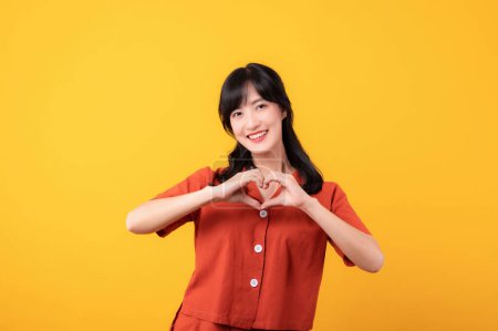 Foto de Retrato hermosa joven asiática mujer feliz sonrisa vestida en naranja ropa mostrando el corazón gesto de la mano aislado en amarillo estudio fondo. - Imagen libre de derechos