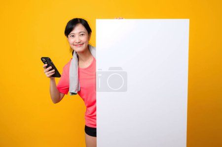 Photo for Asian young sports fitness woman happy smile wearing pink sportswear standing behind the white blank banner or empty space advertisement board against yellow background. wellbeing application concept. - Royalty Free Image