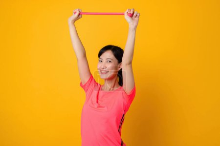 Téléchargez les photos : Portrait asiatique jeune femme de fitness sportive heureuse de porter des vêtements de sport roses et bande de résistance étirant faire de l'exercice séance d'entraînement sur fond jaune. bien-être mode de vie sain concept. - en image libre de droit