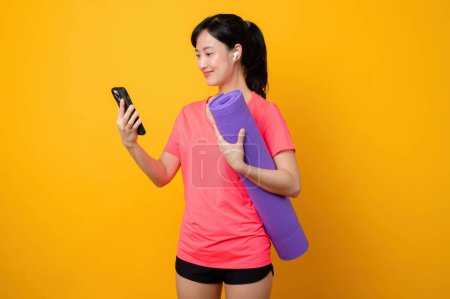 Foto de Retrato asiático joven deportista fitness mujer sonrisa feliz usando ropa deportiva rosa y teléfono inteligente haciendo ejercicio de entrenamiento con fondo de estudio amarillo. tecnología bienestar estilo de vida concepto. - Imagen libre de derechos