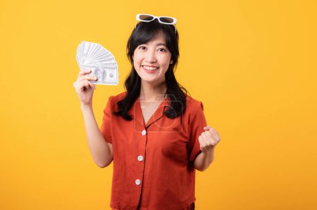 Foto de Retrato hermosa joven asiática mujer feliz sonrisa vestida con ropa naranja sosteniendo dinero en efectivo y celebrar su éxito aislado sobre fondo amarillo. Ser rico concepto. - Imagen libre de derechos