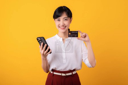 Photo for Portrait young beautiful asian woman enterpriser happy smile wearing white shirt and red plants holding credit card and smartphone isolation on yellow background. Online shopping concept. - Royalty Free Image