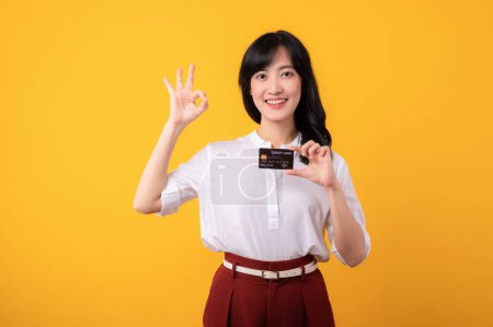 Photo for Portrait young beautiful asian woman enterpriser happy smile wearing white shirt and red plants showing okay hand gesture and holding credit card isolation on yellow background. - Royalty Free Image