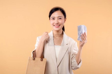 Photo for Portrait of successful happy confident young asian business woman wearing white jacket holding cash money dollars and eco bag standing over beige background. Eco friendly shopping concept. - Royalty Free Image
