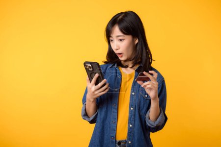 Photo for Portrait beautiful asian young woman happy smile dressed in denim jacket showing smartphone and credit card isolate on yellow studio background. Shopping online smartphone application concept - Royalty Free Image