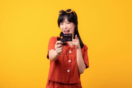 Téléchargez les photos : Portrait belle jeune femme asiatique sourire heureux habillé en vêtements orange montrant carte de crédit et pointeur vers la caméra isolée sur fond jaune. Paiement et achat shopping concept de paiement. - en image libre de droit