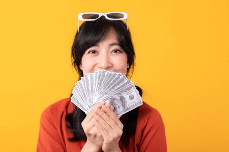 Téléchargez les photos : Portrait belle jeune femme asiatique sourire heureux habillé en vêtements orange montrant argent comptant isolé sur fond jaune. Paiement et achat shopping concept de paiement. - en image libre de droit