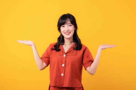 Photo for Portrait beautiful young asian woman happy smile dressed in orange clothes showing hand gesture, arm compare two variants demonstrate empty space isolated on yellow studio background. - Royalty Free Image