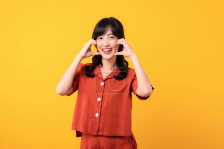 Téléchargez les photos : Portrait belle jeune femme asiatique sourire heureux habillé en vêtements orange montrant les mains du cœur toucher geste joue isolé sur fond de studio jaune. - en image libre de droit