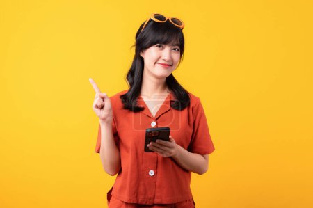 Téléchargez les photos : Portrait jeune belle femme asiatique heureux sourire vêtu de vêtements orange avec smartphone et pointant doigt geste de la main pour libérer de l'espace isolé sur fond de studio jaune. app smartphone concept - en image libre de droit