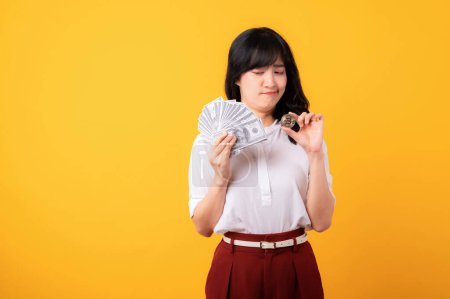 Photo for Portrait young beautiful asian woman enterpriser with doubt face wearing white shirt and red plants holding cash dollar money and crypto digital currency isolation on yellow background. Wealth concept - Royalty Free Image